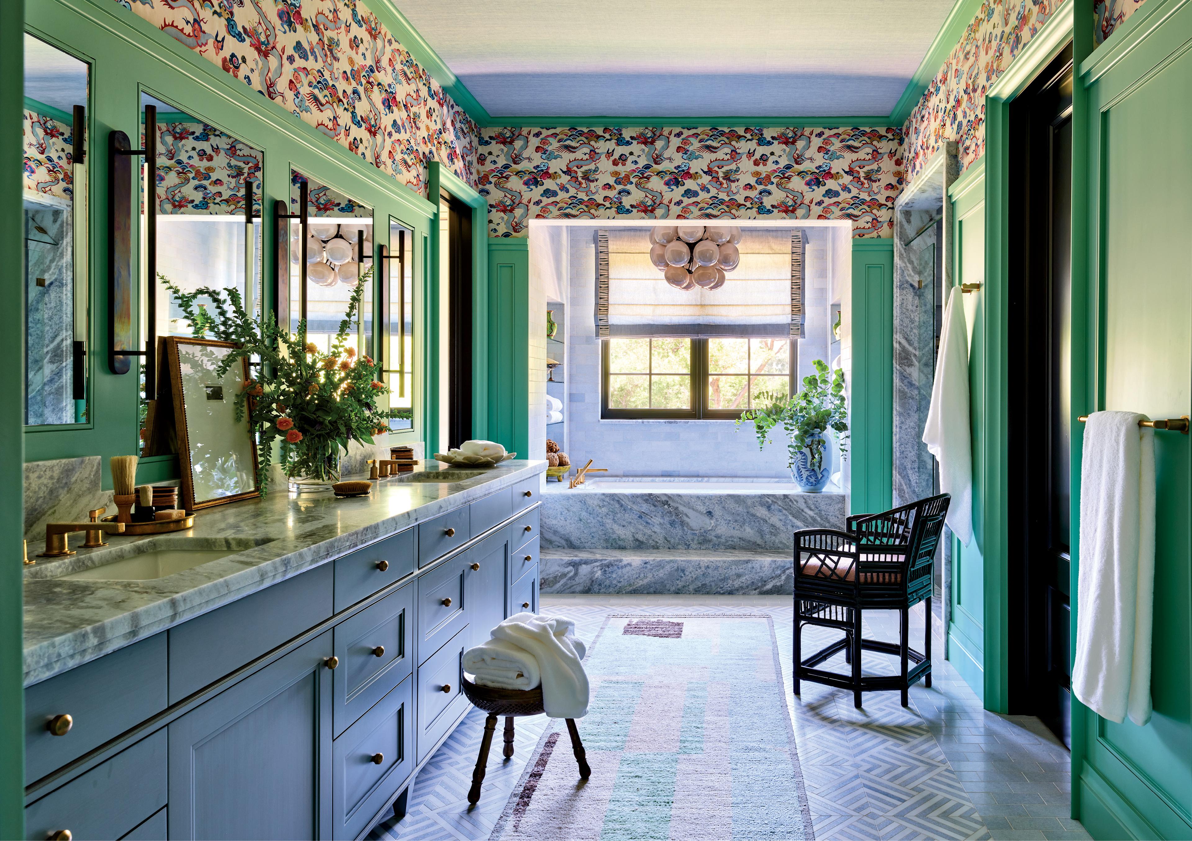 Elegant bathroom with vibrant wallpaper, green trim, and blue cabinetry. Features double sinks, a large mirror, marble surfaces, and a sunlit tub.