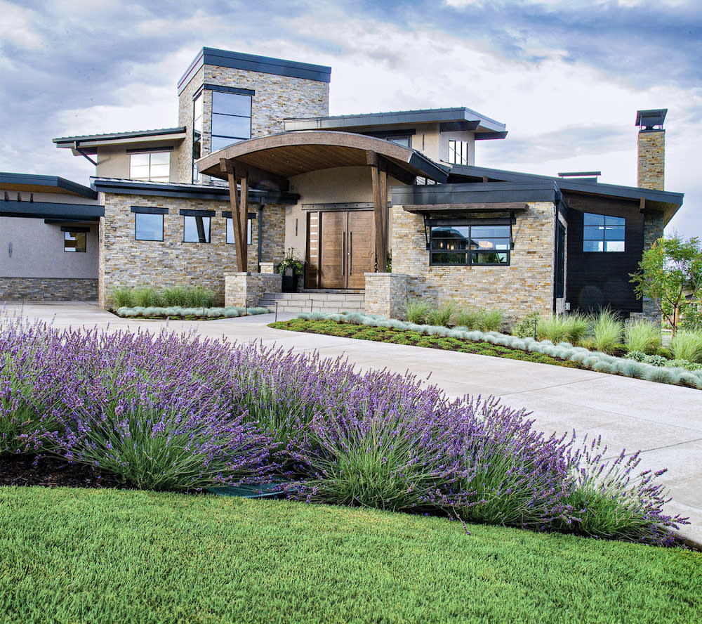 A spacious house with a wide driveway and a beautiful flower bed in the front, adorned with purple flowers and lush green grasses.