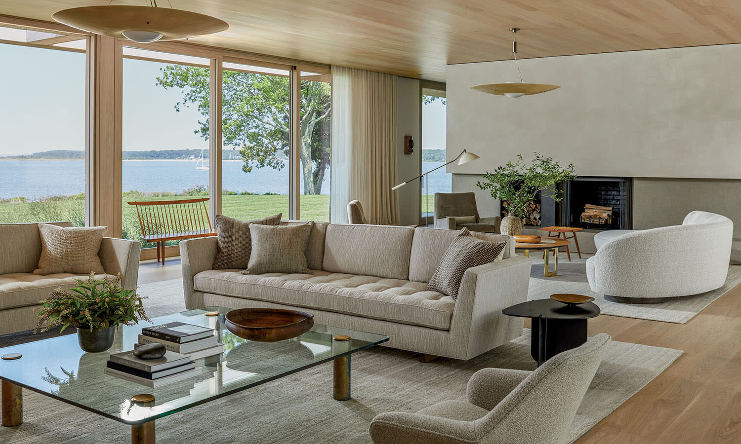 Stylish, modern living room with beige sofas, glass coffee table, and large windows offering a serene view of a lake and greenery. Cozy, inviting atmosphere.