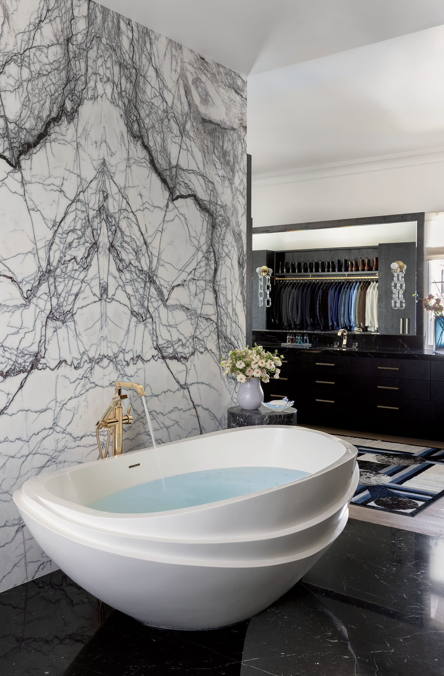 Luxurious bathroom with a white oval tub on a dark marble floor. A striking black and white marble wall adds elegance. A modern sink and organized closet are in view.