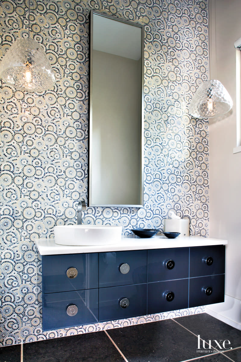 A powder room features an elaborate mosaic tile lines one wall. A blue vanity has a tall mirror above. Two pendants hang on either side of the mirror.