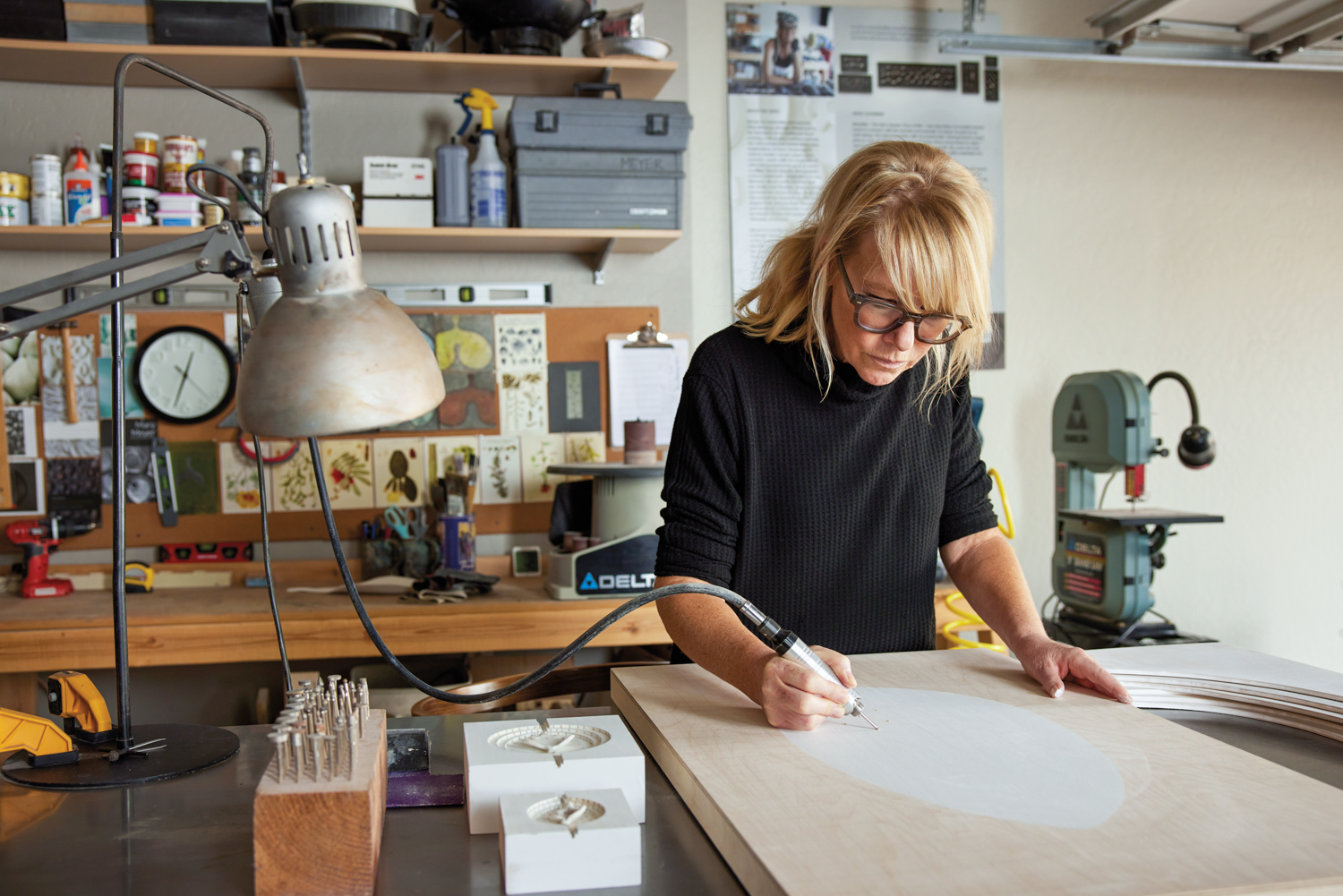 An blonde woman using an electric shaft tool to carve on a wood panel.