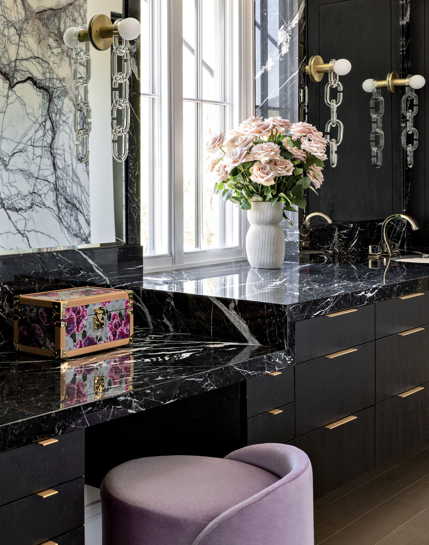 Luxurious bathroom with black marble countertops, gold fixtures, and a vase of pink flowers. A lilac stool adds a touch of elegance and color.