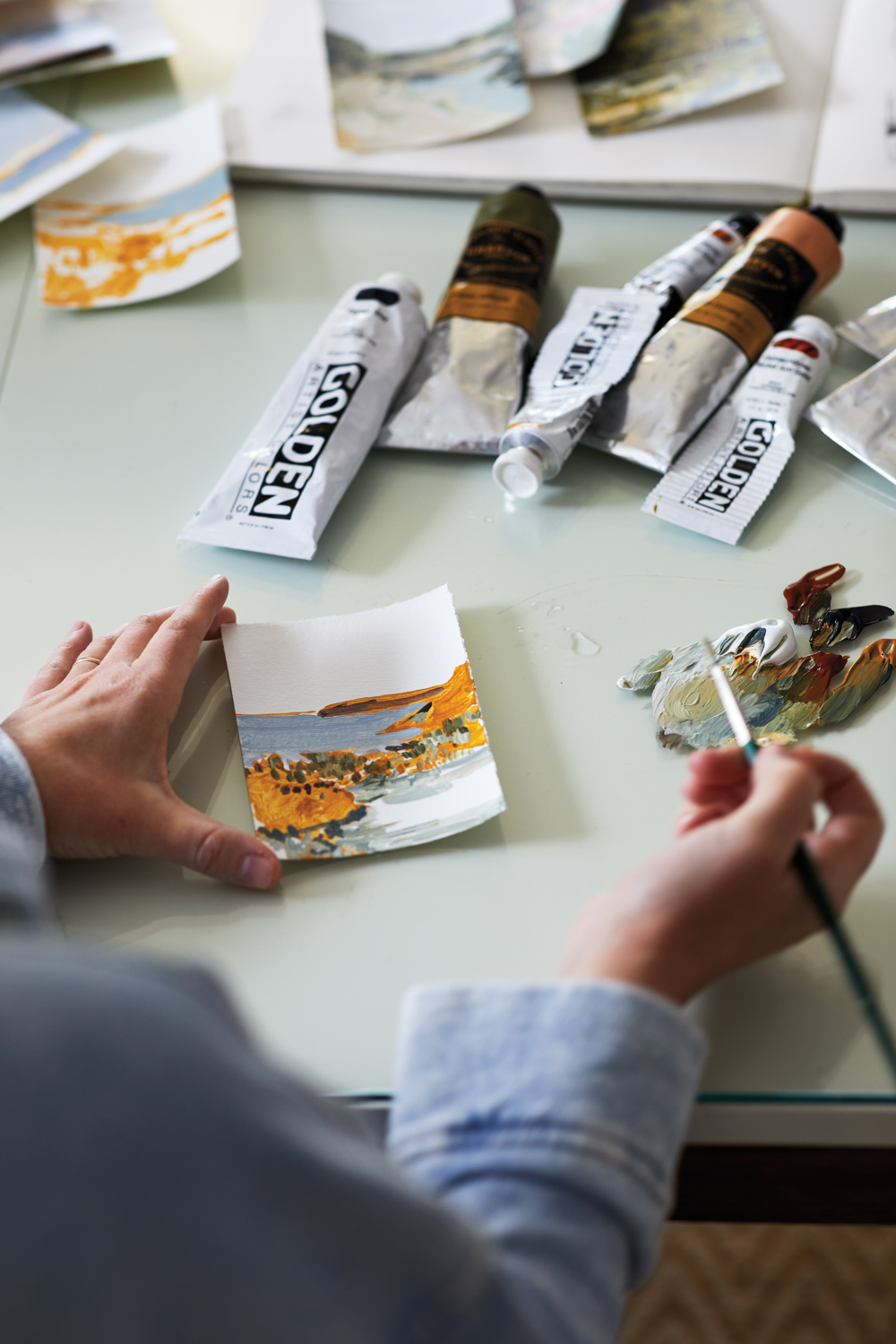 hand holding a paintbrush above a small painting and tubes of pigment