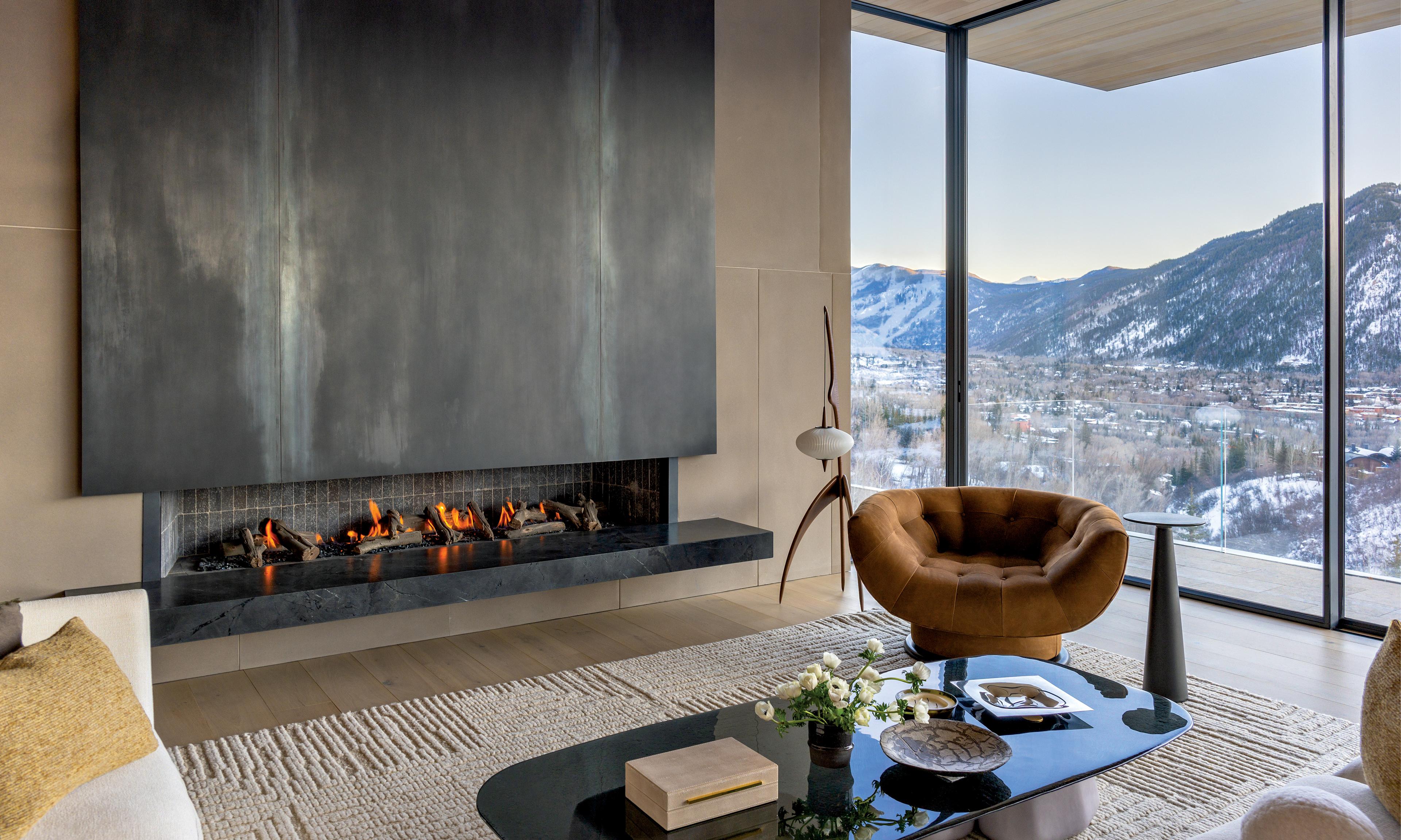 Modern living room with large black fireplace, brown lounge chair, and glass table. Floor-to-ceiling windows reveal snowy mountain views, creating a cozy and serene atmosphere.