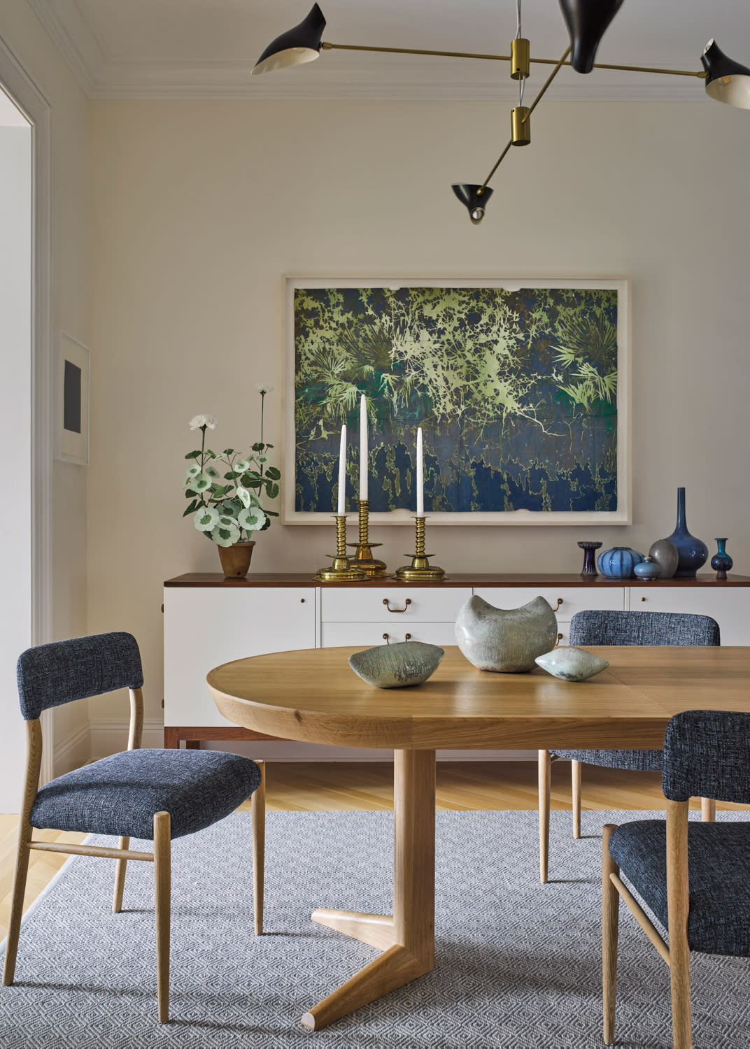 Elegant dining room with a wooden oval table, blue upholstered chairs, and ceramic bowls. A large plant artwork and candle holders adorn the sideboard.