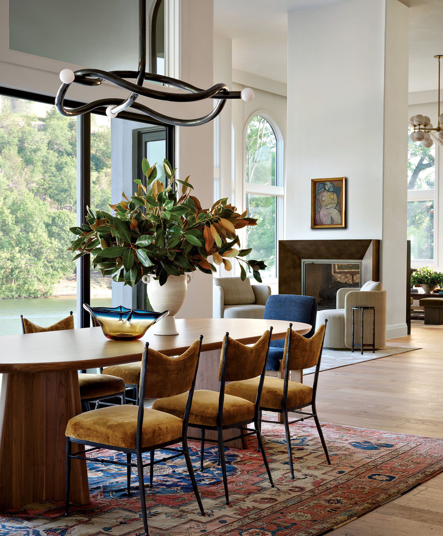 A stylish kitchen features dark cabinetry, a marble backsplash, and elegant shelves. Brown leather chairs surround a marble island topped with fresh produce and a large plant, under a glass chandelier.