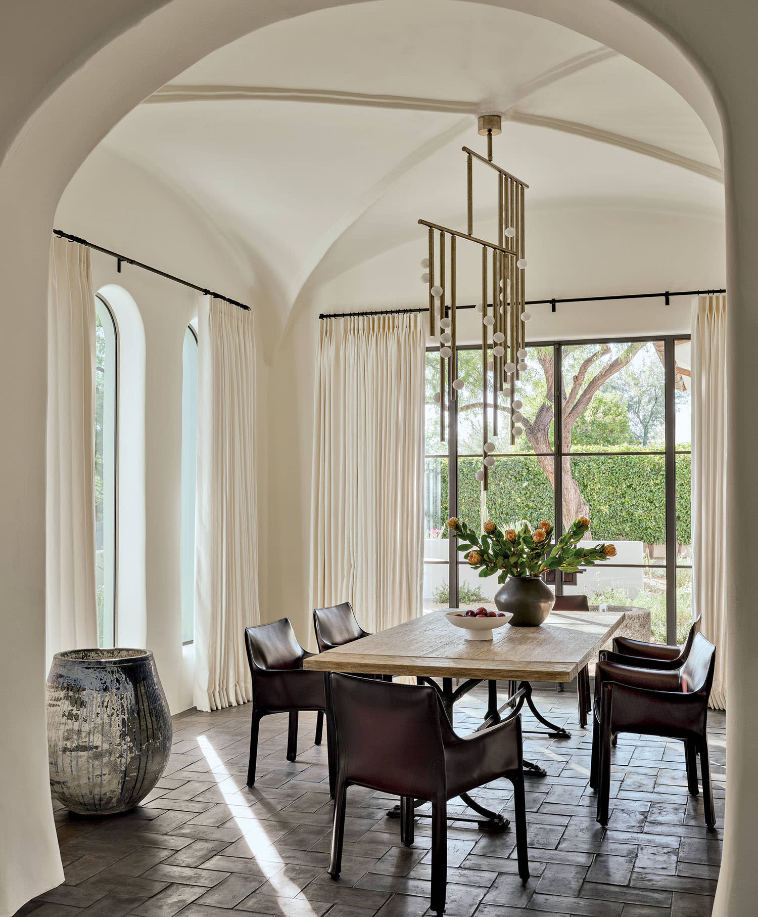 A chandelier hangs in a dining room set with a table and six chairs in front of a large window.