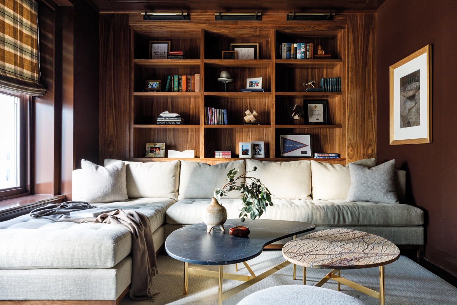 Creamy white sectional in front of wood shelving and next to windows with plaid Roman shades