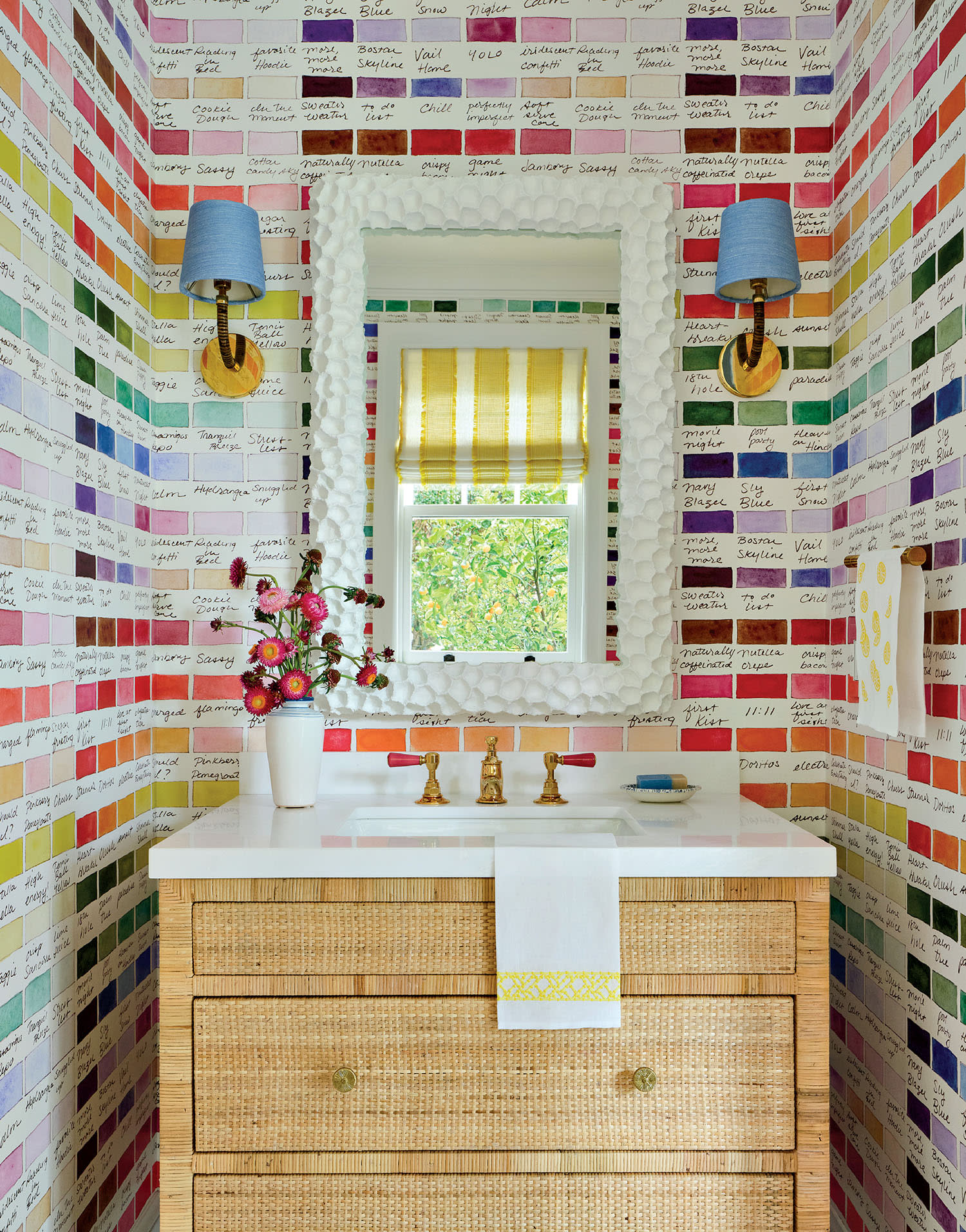 Colorful bathroom with walls covered in multicolored paint swatch samples. A wicker vanity with brass faucet, blue wall sconces, and a vase of flowers add charm.