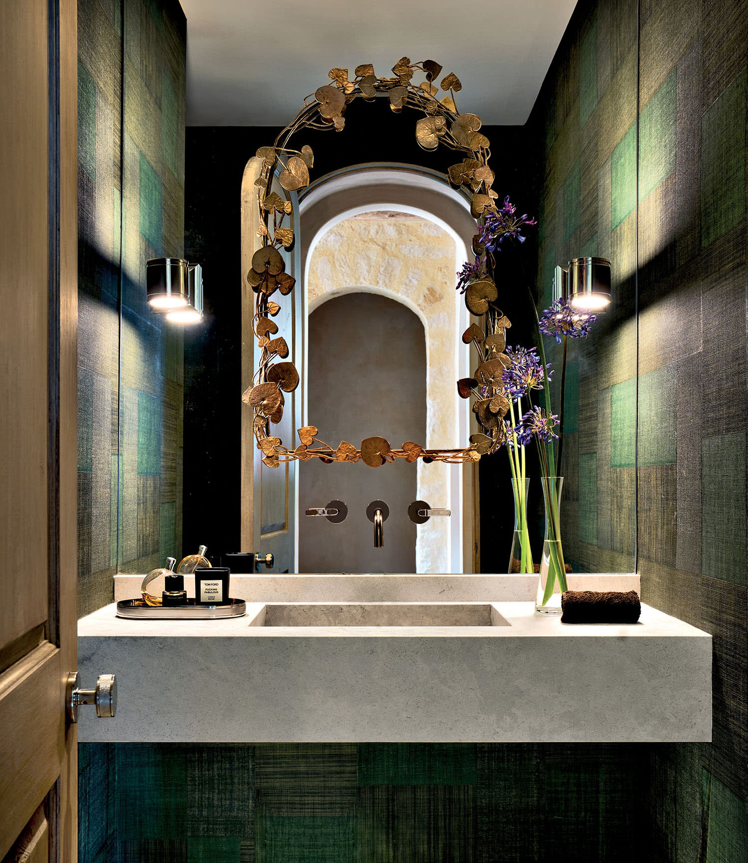 Stylish bathroom with dark green textured walls, a unique oval mirror with decorative frame, built-in sink, modern sconces, and purple flowers in a vase.