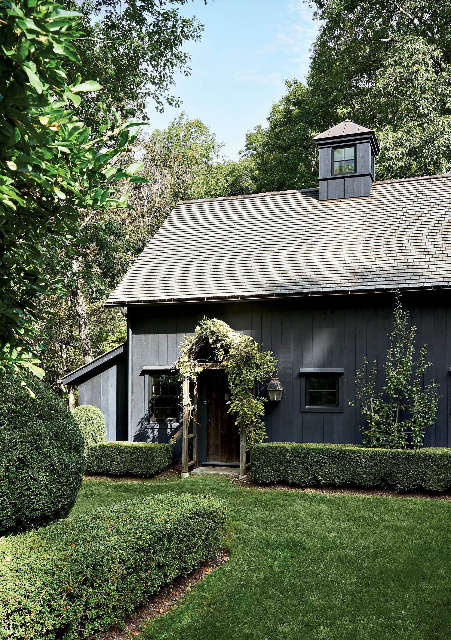 A barn with a dark gray exterior sits on a green lawn with nearby trees.