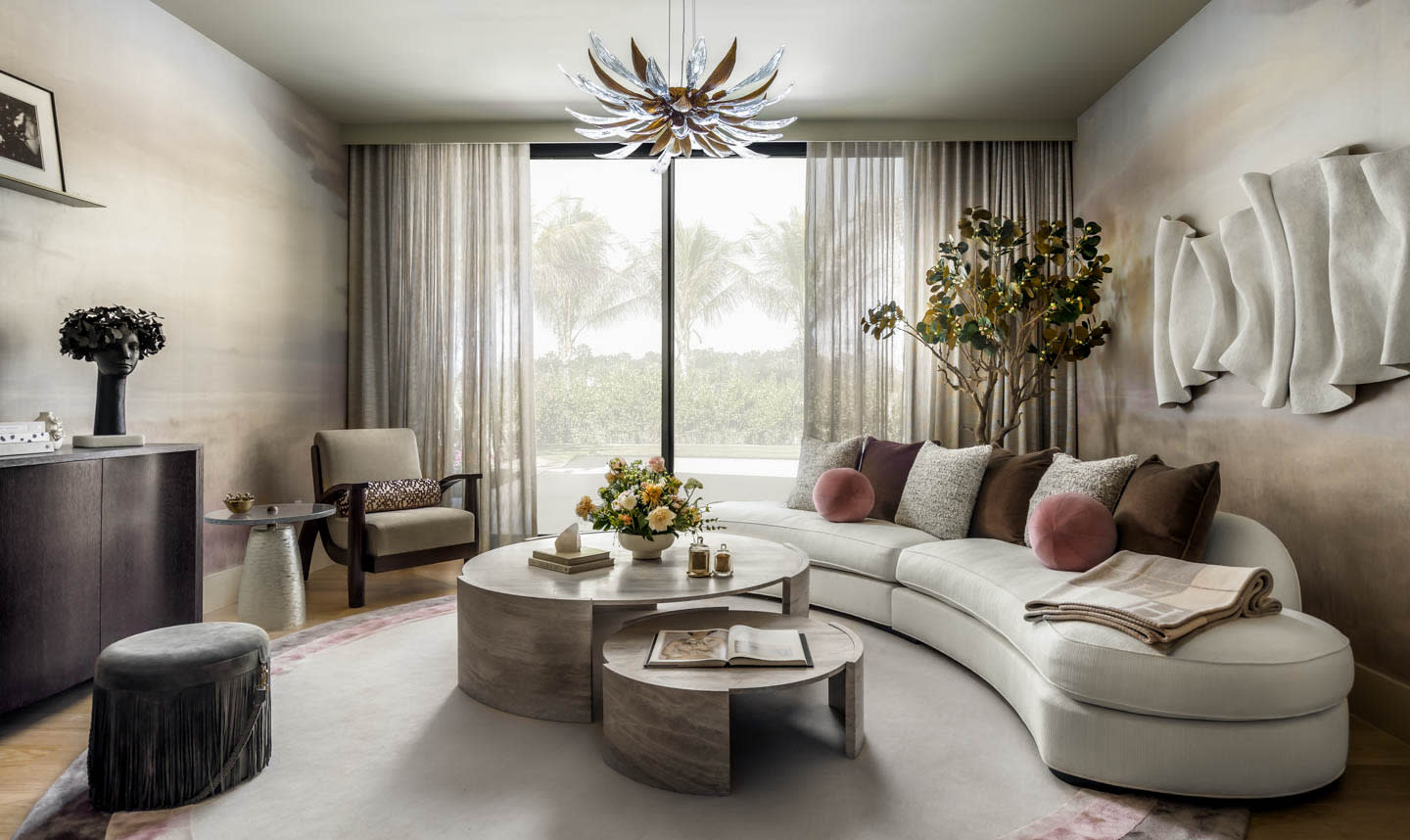 Luxurious living room with a curved white sofa adorned with cushions, circular coffee tables, large window with sheer curtains, modern chandelier, and wall art.
