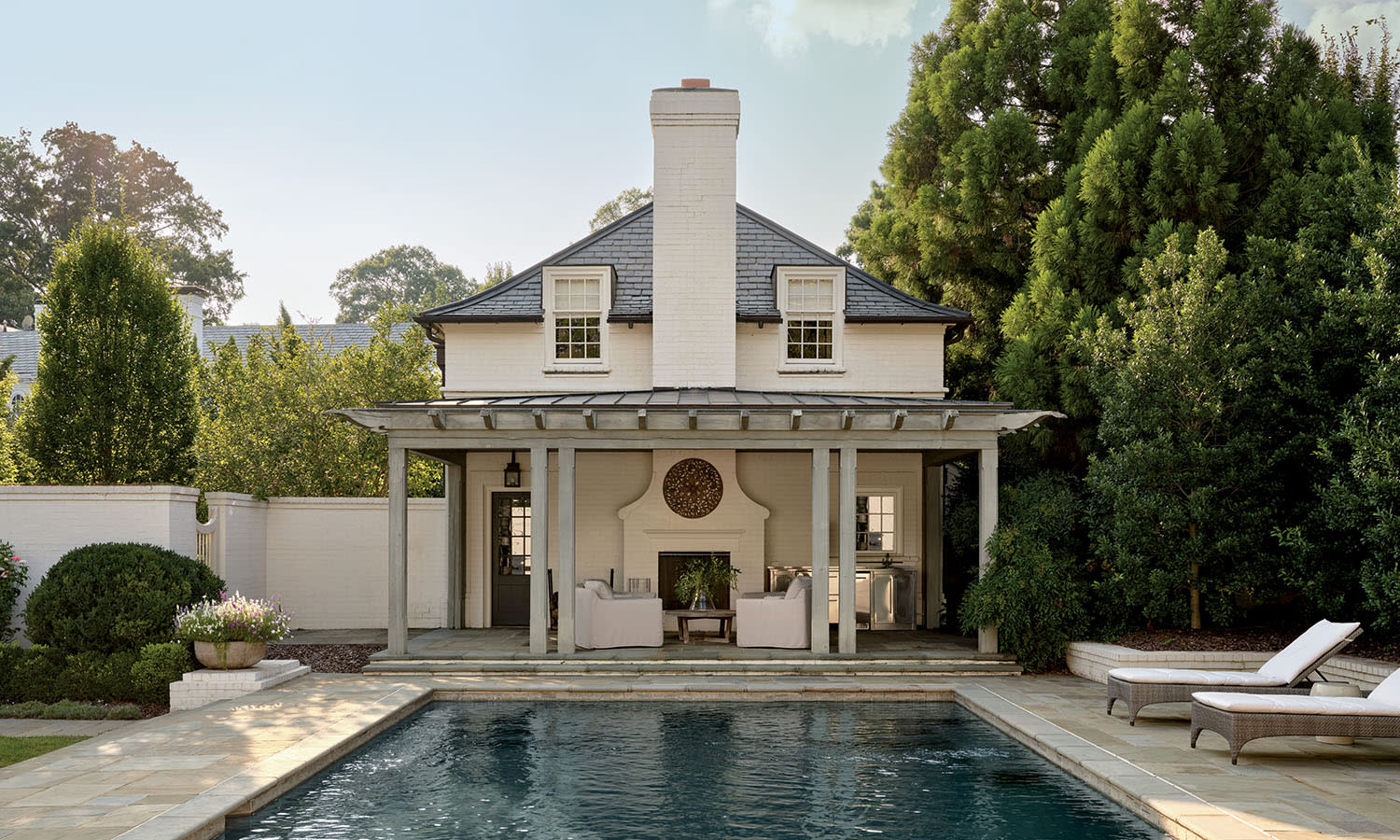 A pool and patio area in front of a white house, featuring lounge chairs and greenery surrounding the space.