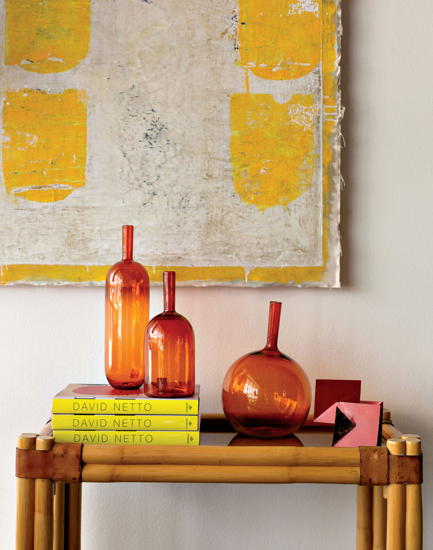 Orange glass bottles sit on a stack of yellow books with a white-and-yellow painting hung above.