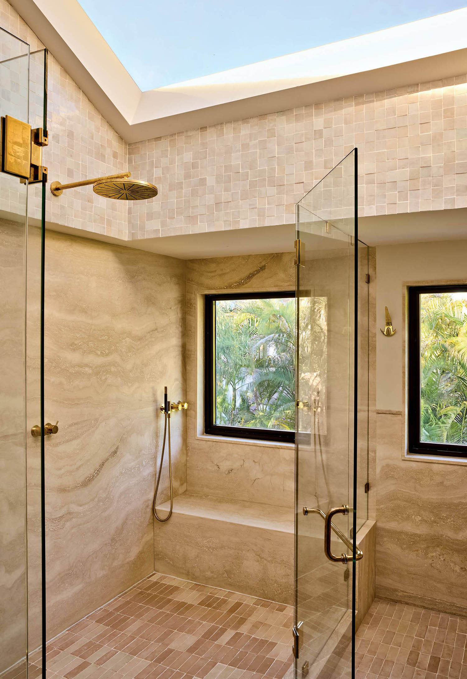 A large skylight and windows let light into a stone shower constructed with benches and glass doors.