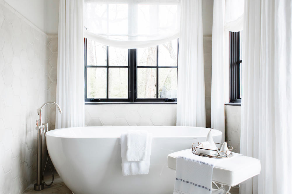 Stone hex tiles from floor to ceiling and a white porcelain tub are a vision of softness amongst the floor-to-ceiling sheers and relaxed window valances.