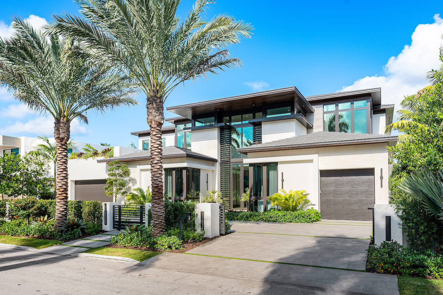 Contemporary two-story coastal home with cream stucco walls, slate-gray rooflines, and a dramatic glass entry tower fronted by slender vertical windows. Twin palm trees shade a gated driveway bordered by lush hedges, tropical ferns, and manicured lawns under a vivid blue, cloud-dotted sky.