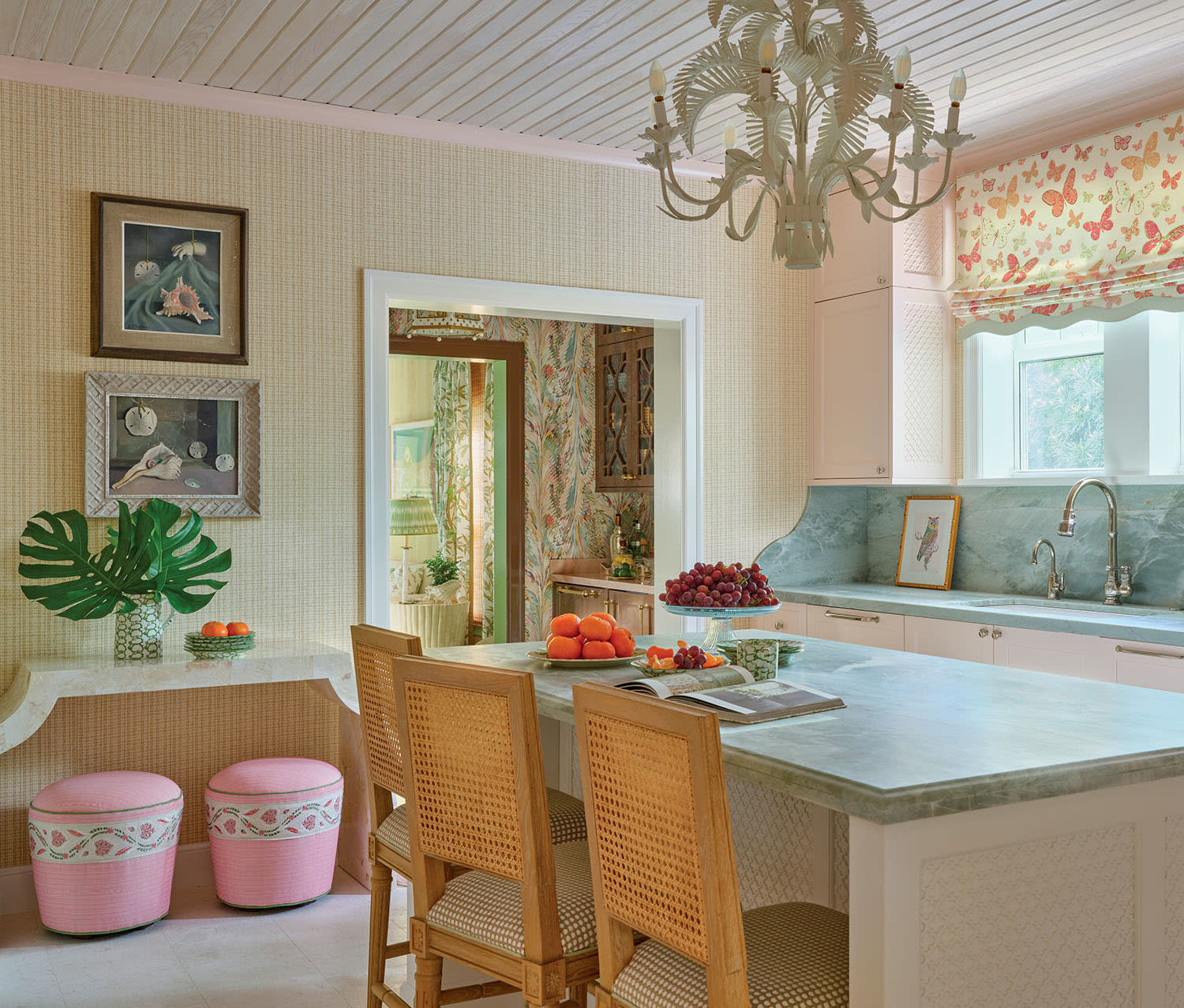 Bright kitchen with pastel tones, featuring a green marble island, pink butterfly curtains, elegant chandelier, and vibrant fruit bowl centerpiece.