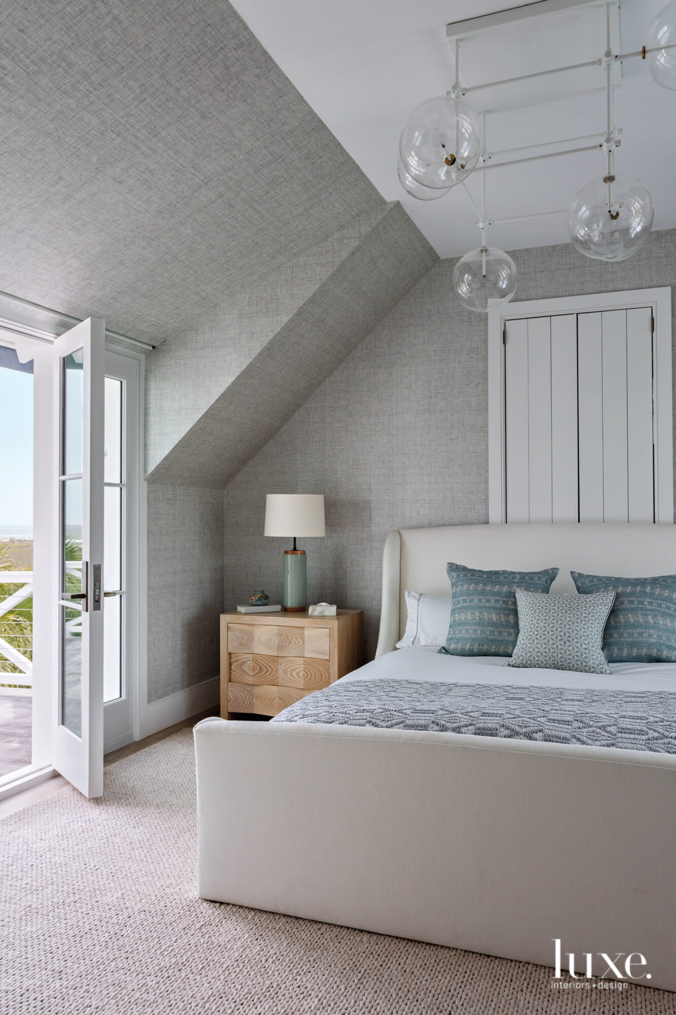 Bedroom with gross-hatched gray walls, ivory upholstered bed, teal pillows, gray blanket, clear glass close chandelier, honey-colored nightstand, French doors to porch