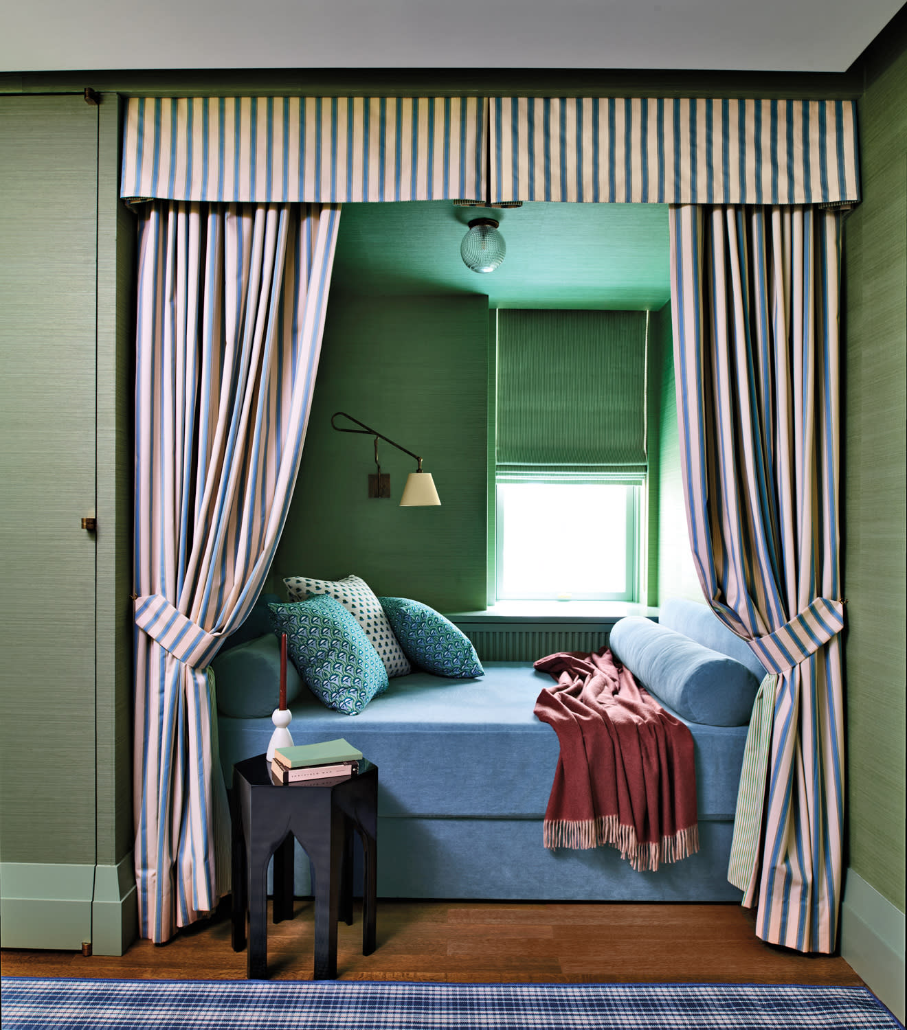 Cozy nook with a blue daybed surrounded by striped curtains. It features patterned pillows, a red throw, and a small black side table with books.