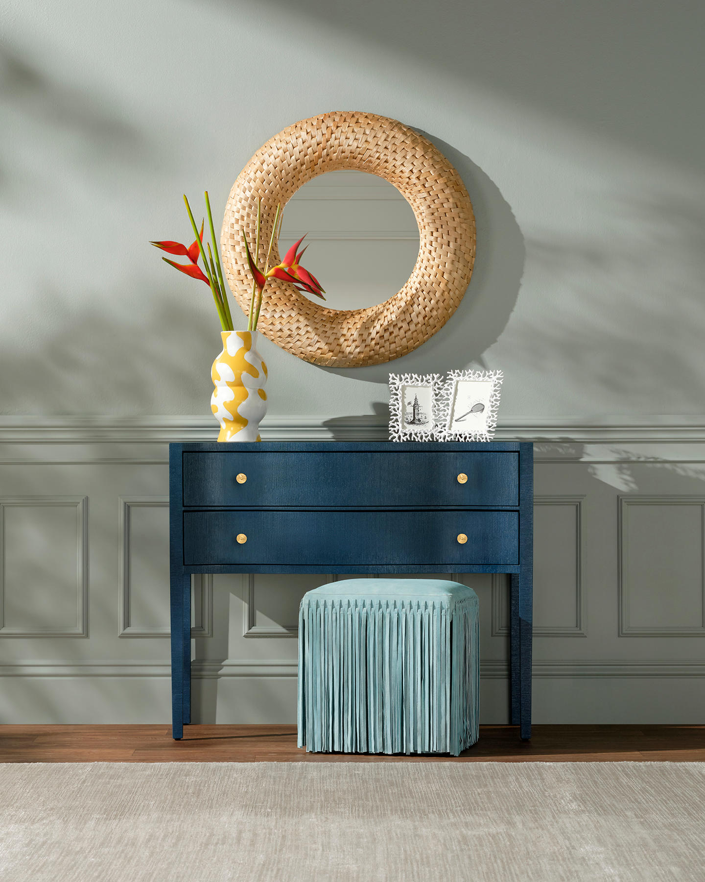 A navy console with gold knobs against a light gray wall features a round woven mirror above. A yellow vase with orange flowers and framed photos adorn the top, with a blue fringed stool below. The scene is calm and stylish with soft shadows.