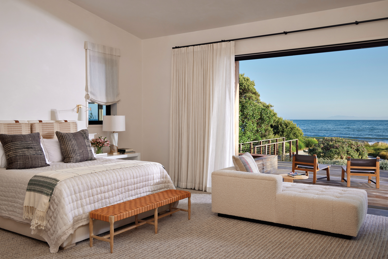 bedroom with neutral fabrics, woven bench, and chaise lounge facing deck area with two sling chairs