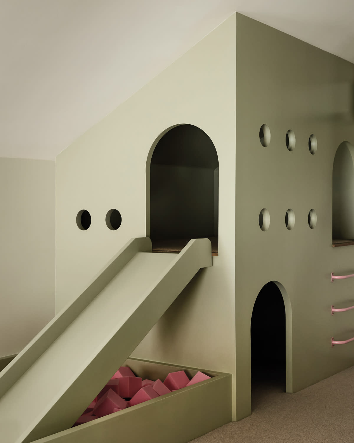 Minimalist playroom with a light green structure featuring a slide, round openings and climbing holds
