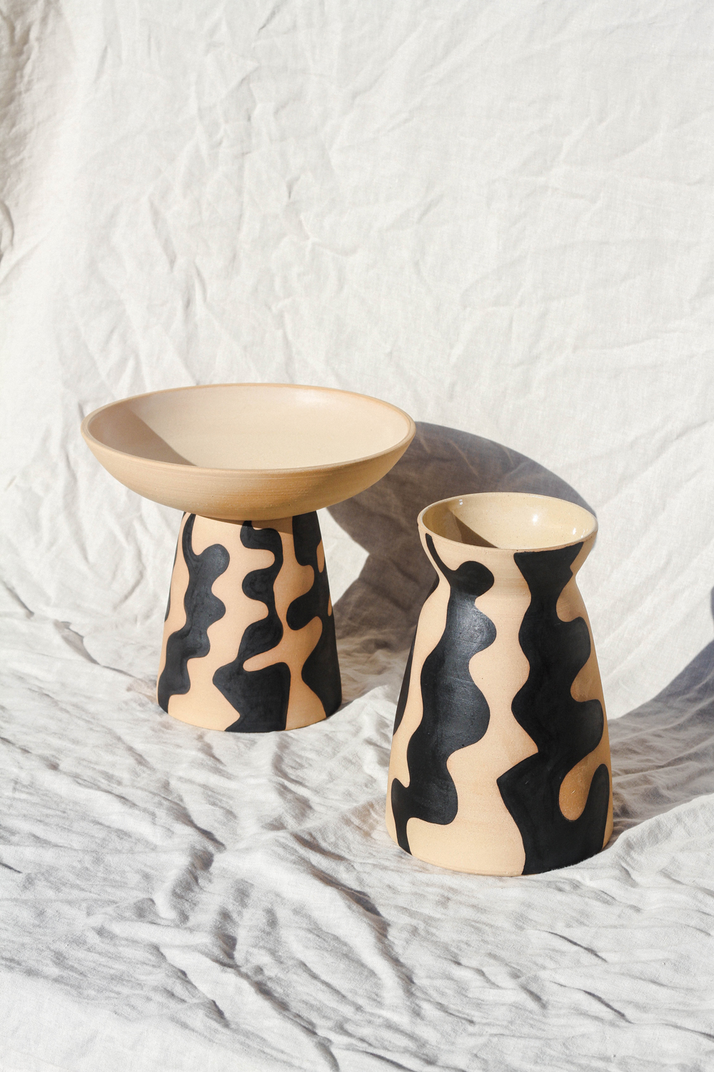 2 cream and black ceramic vessels sitting on a white cloth backdrop.