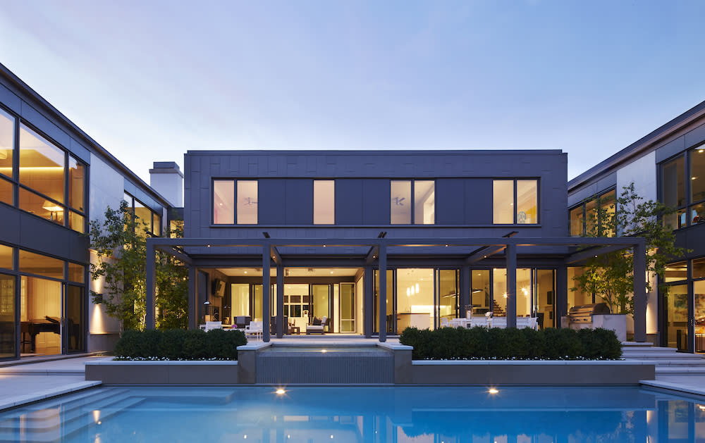 Exterior of home at dusk with lights illuminating the inside and the large pool that is in the center of yard.