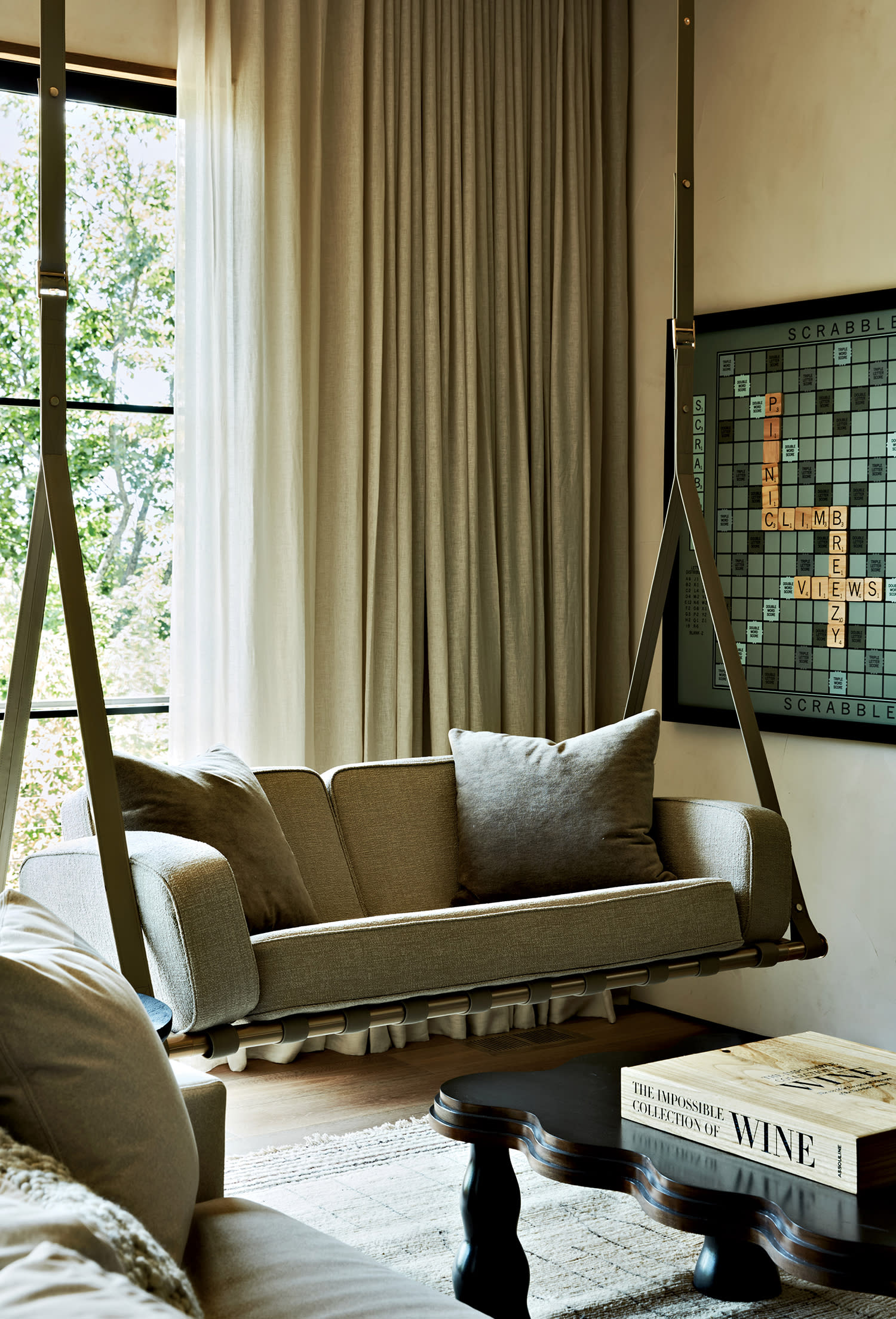 A cushioned seat hangs near a window, set above a white wool rug with a dark coffee table on top of it.  