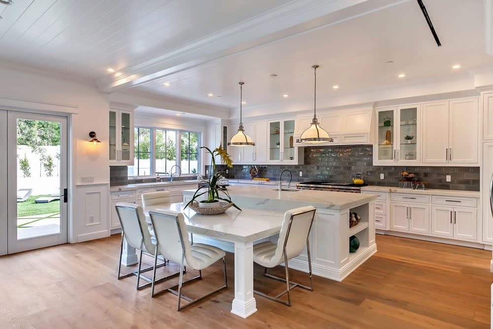 A modern white kitchen featuring a dining table surrounded by chairs and a black tile backsplash.