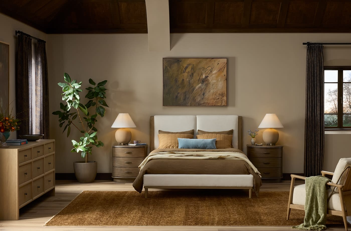 Cozy bedroom with a neutral palette, featuring a bed with brown and beige bedding, flanked by wooden nightstands with lamps. A large plant and abstract art add warmth.