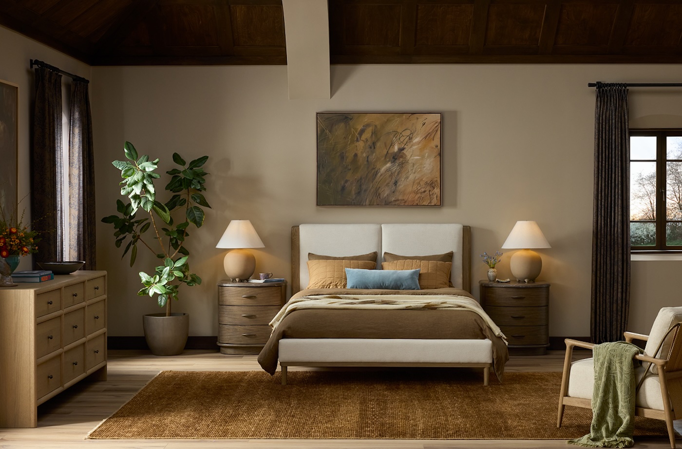 Cozy bedroom with a neutral palette, featuring a bed with brown and beige bedding, flanked by wooden nightstands with lamps. A large plant and abstract art add warmth.
