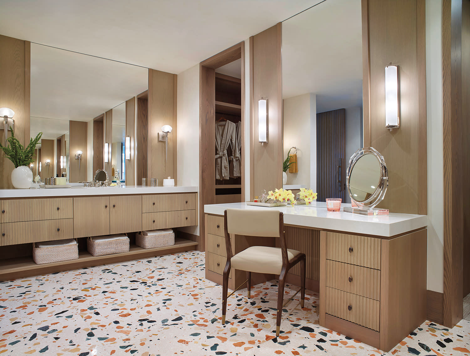 Spacious vanity area with wood cabinetry, terrazzo floor, and large mirror. Soft lighting, fresh flowers, and plush robes create a luxurious, calm ambiance.
