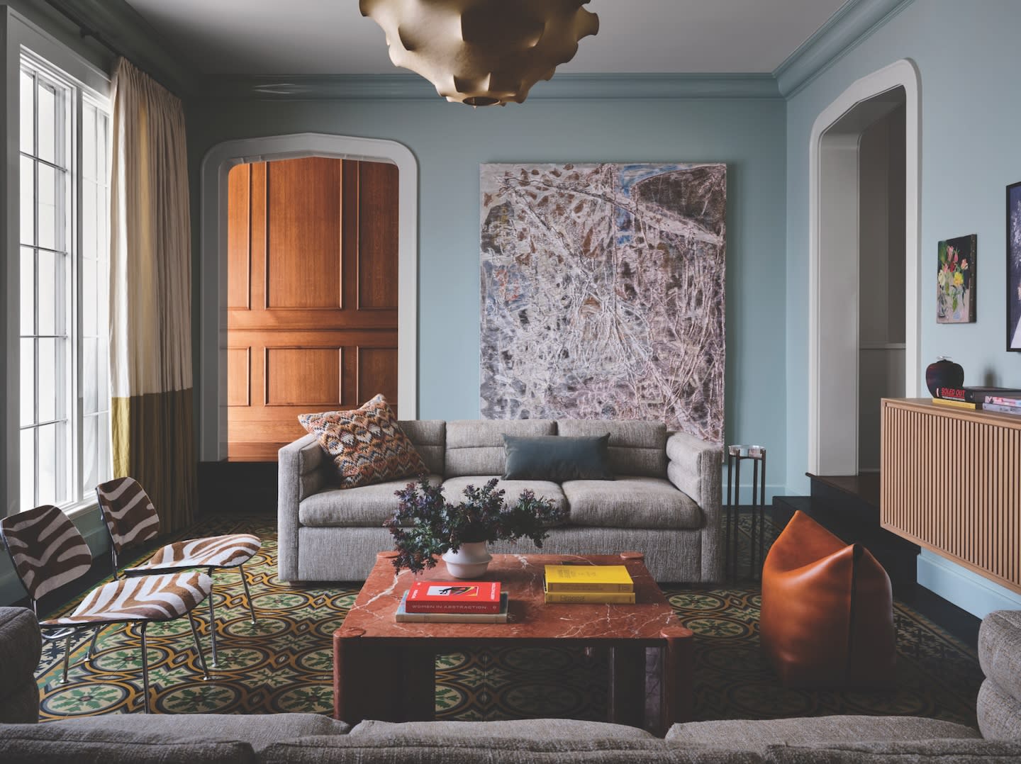 Sophisticated living room with light blue walls, a large gray sofa, and a marble coffee table. Abstract art, patterned chairs, and a cozy atmosphere.