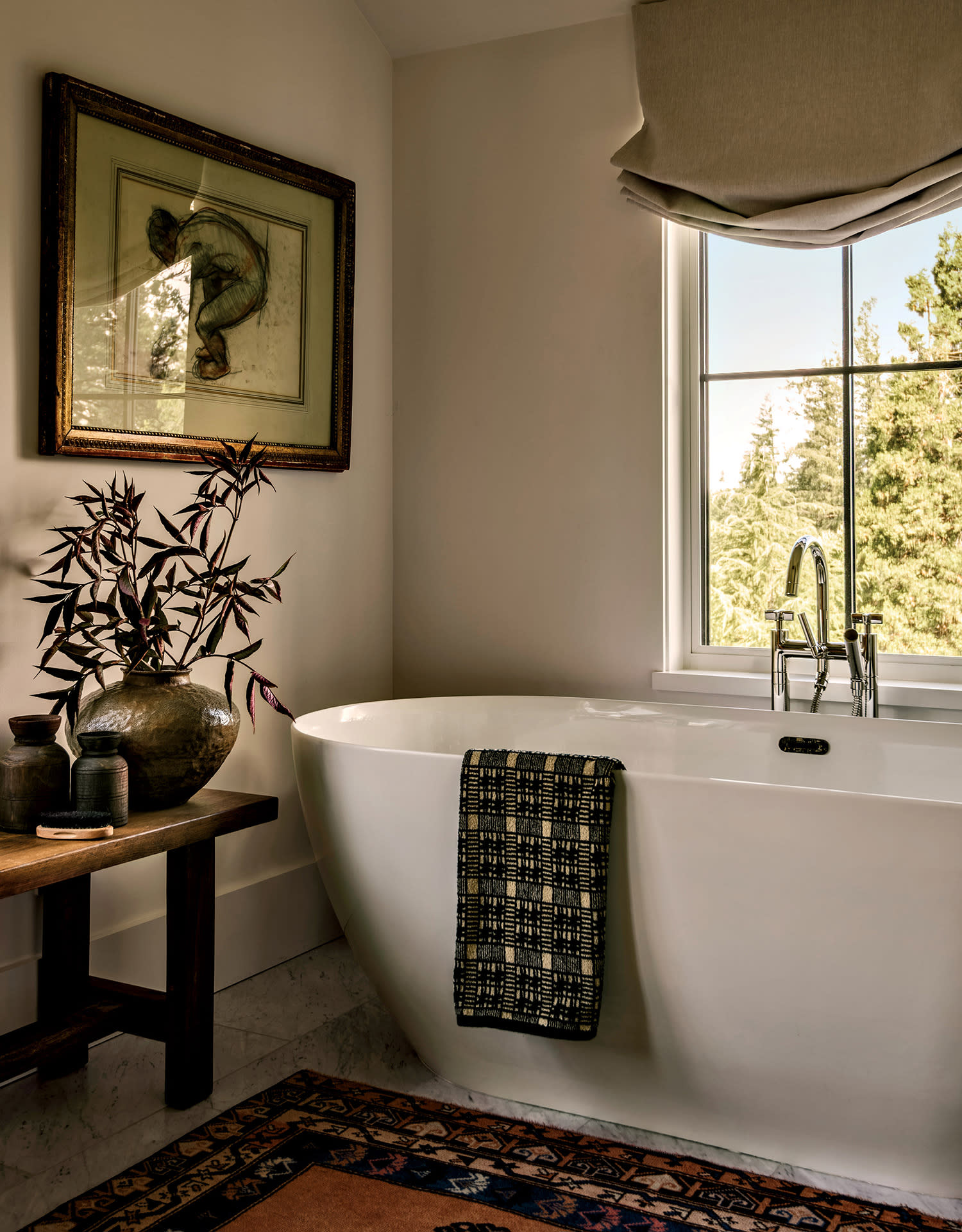 Cozy bathroom with a white freestanding tub, textured towel, and large window with natural light. Decorative vase with plants and framed art add elegance.