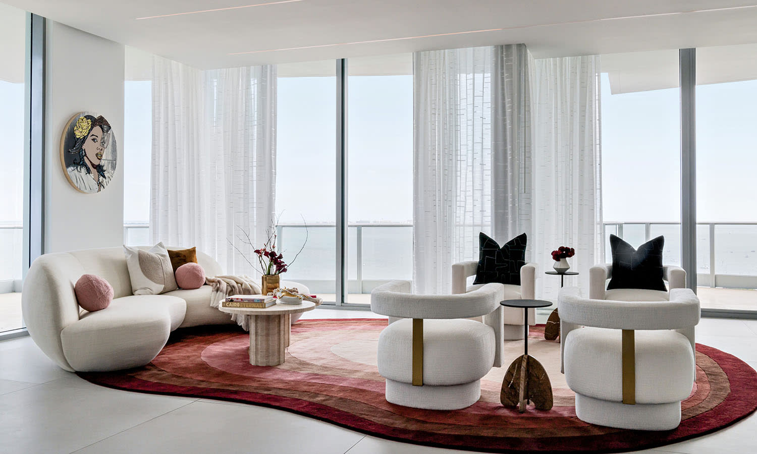 Modern living room with white, curved furniture, a colorful abstract rug, and floor-to-ceiling windows. Soft lighting creates a warm, inviting atmosphere.