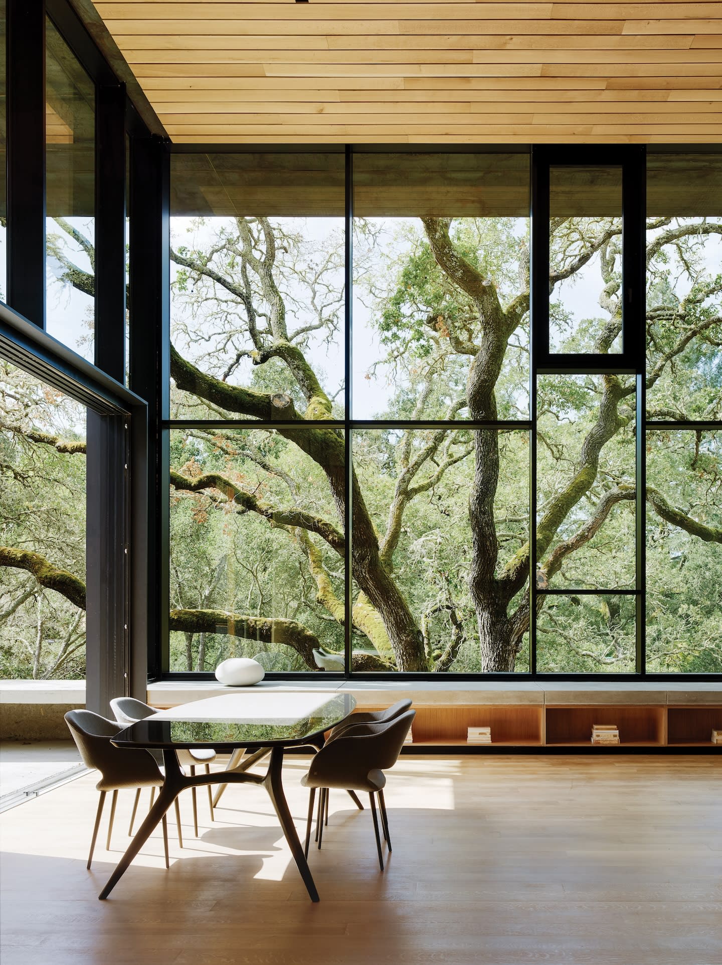 A modern dining room with a glass table and black chairs, set against a large window showcasing lush green trees. The room has a serene, open ambiance.
