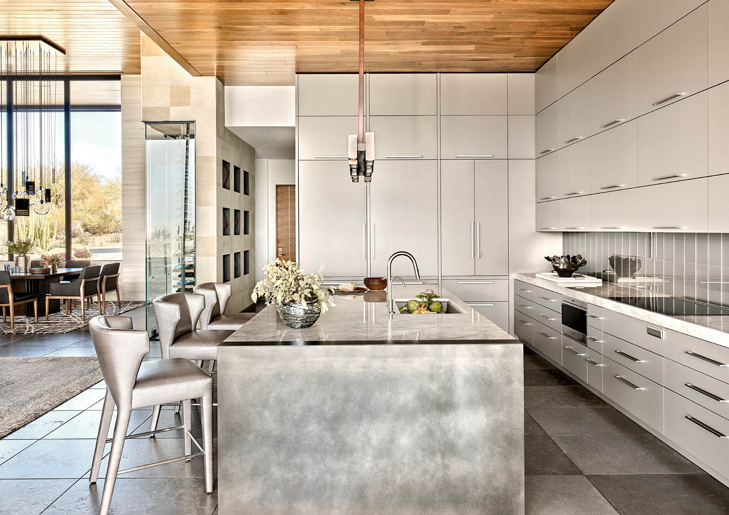 Counter stools sit next to a stone-and-steel island in a kitchen with white cabinets and wood ceilings.