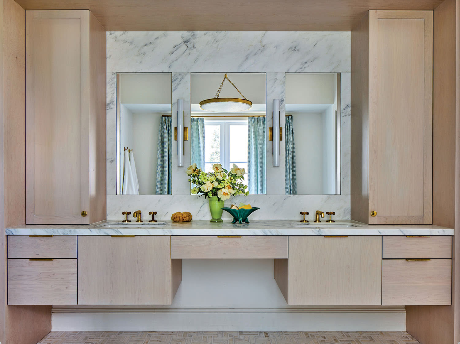 Modern bathroom vanity with marble countertop, dual brass faucets, and two large mirrors. Accented by a hanging light, flowers, and pastel cabinets.