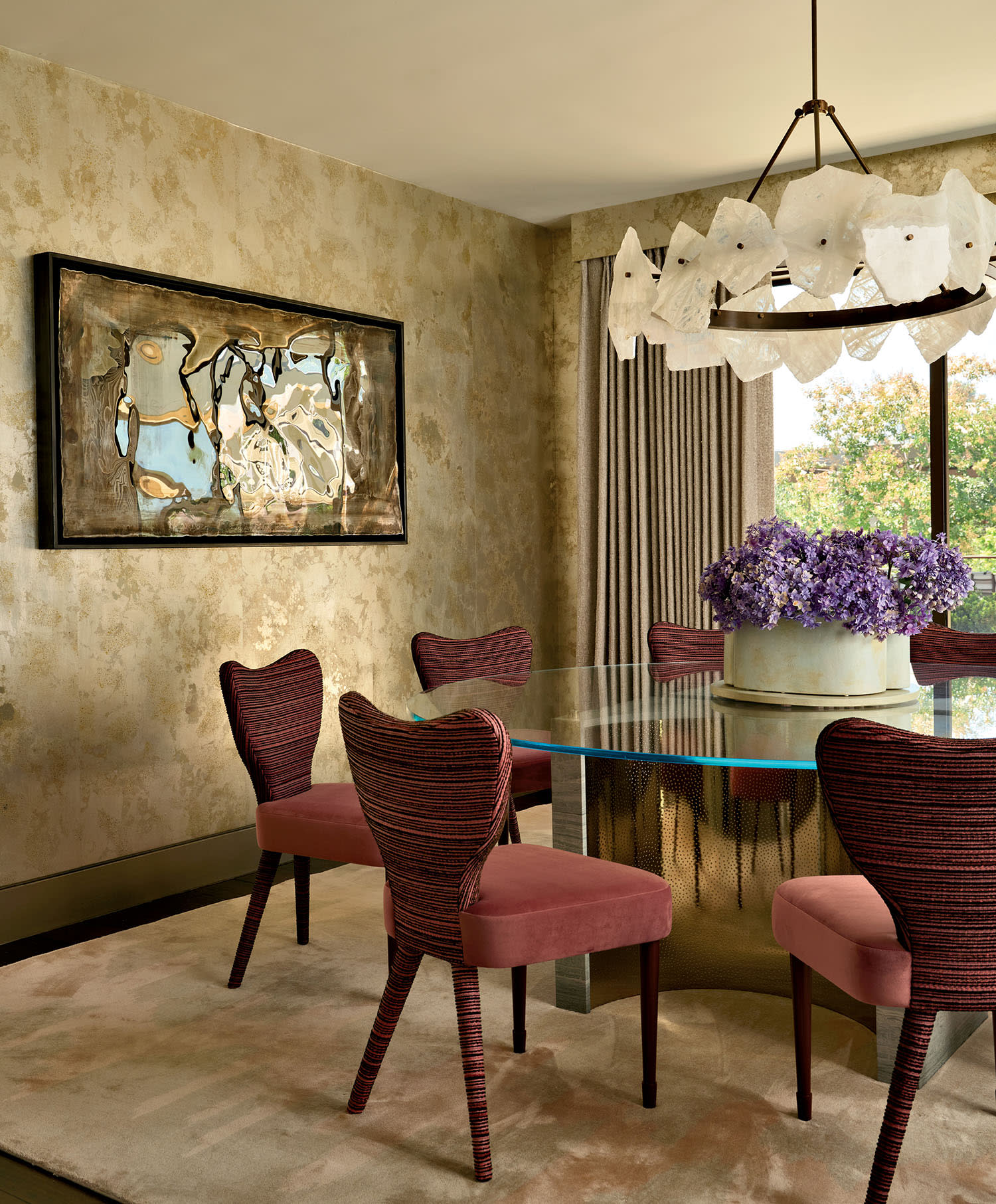 Elegant dining room with a glass table, pink chairs, and a white chandelier. Abstract art on textured wall; a vase of purple flowers adds charm.