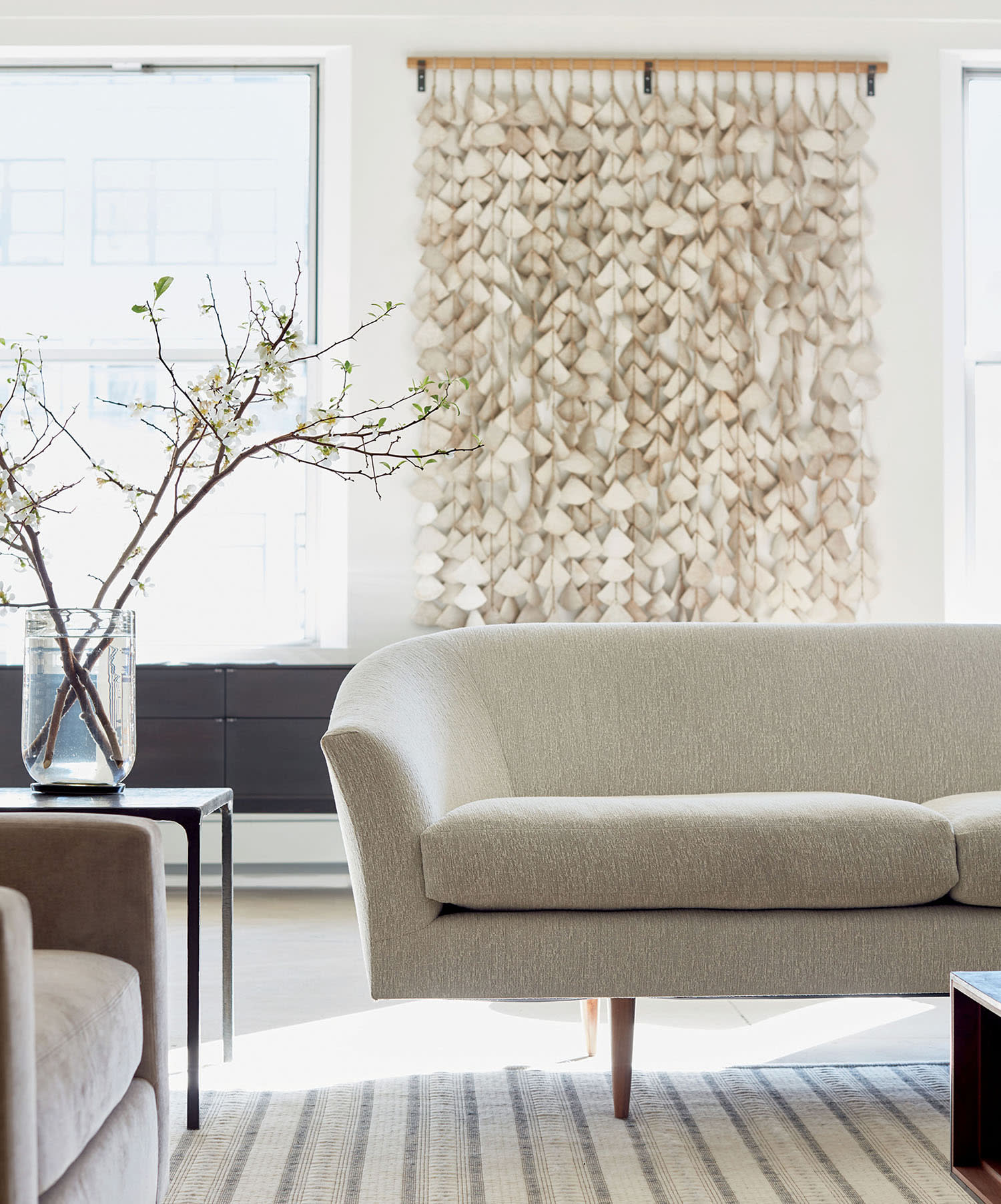 A light-colored couch sits on a rug, near an accent table and decorative wall hanging.