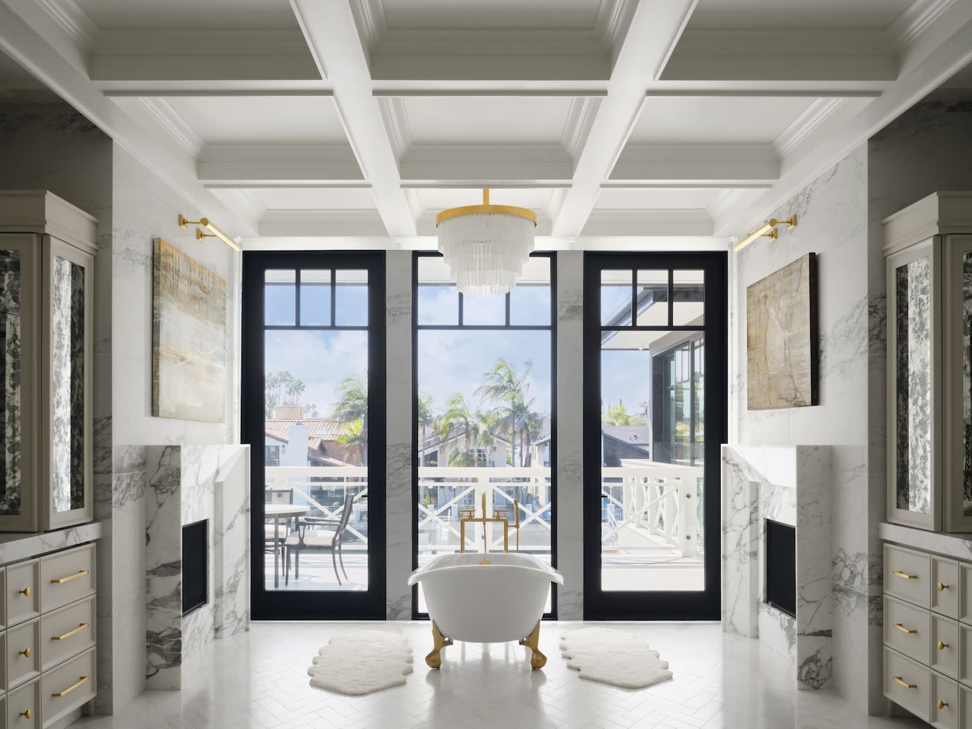 Luxurious bathroom with a freestanding tub, gold fixtures, marble walls, and large windows overlooking palm trees. A chandelier adds elegance.