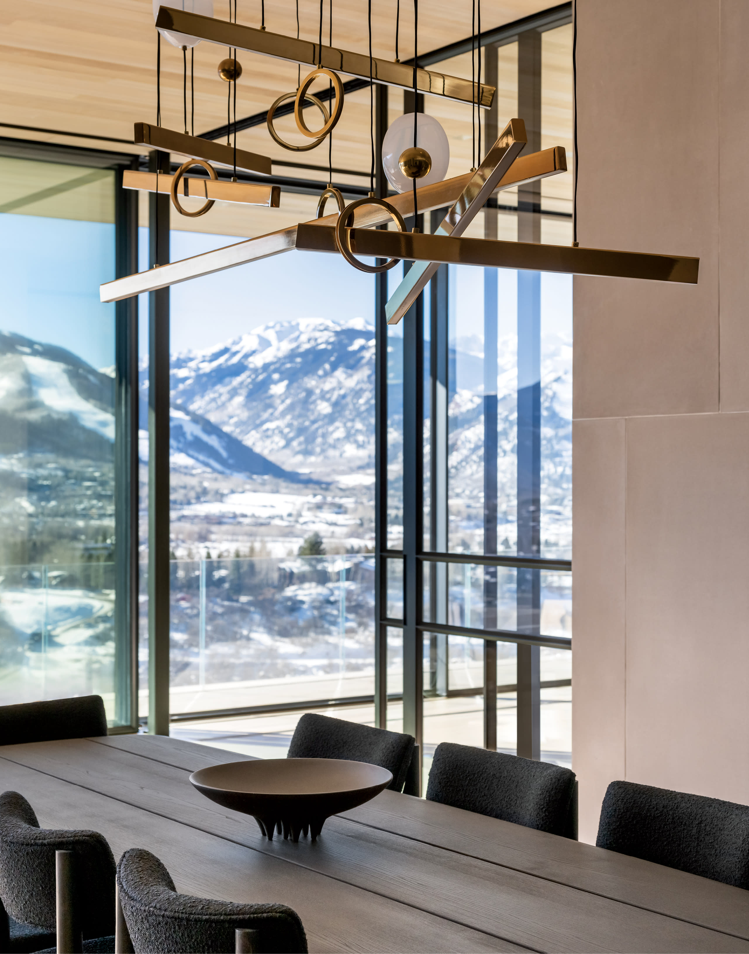 A modern dining room with a long wooden table and dark chairs. An artistic chandelier hangs above. Large windows reveal a snowy mountain view.