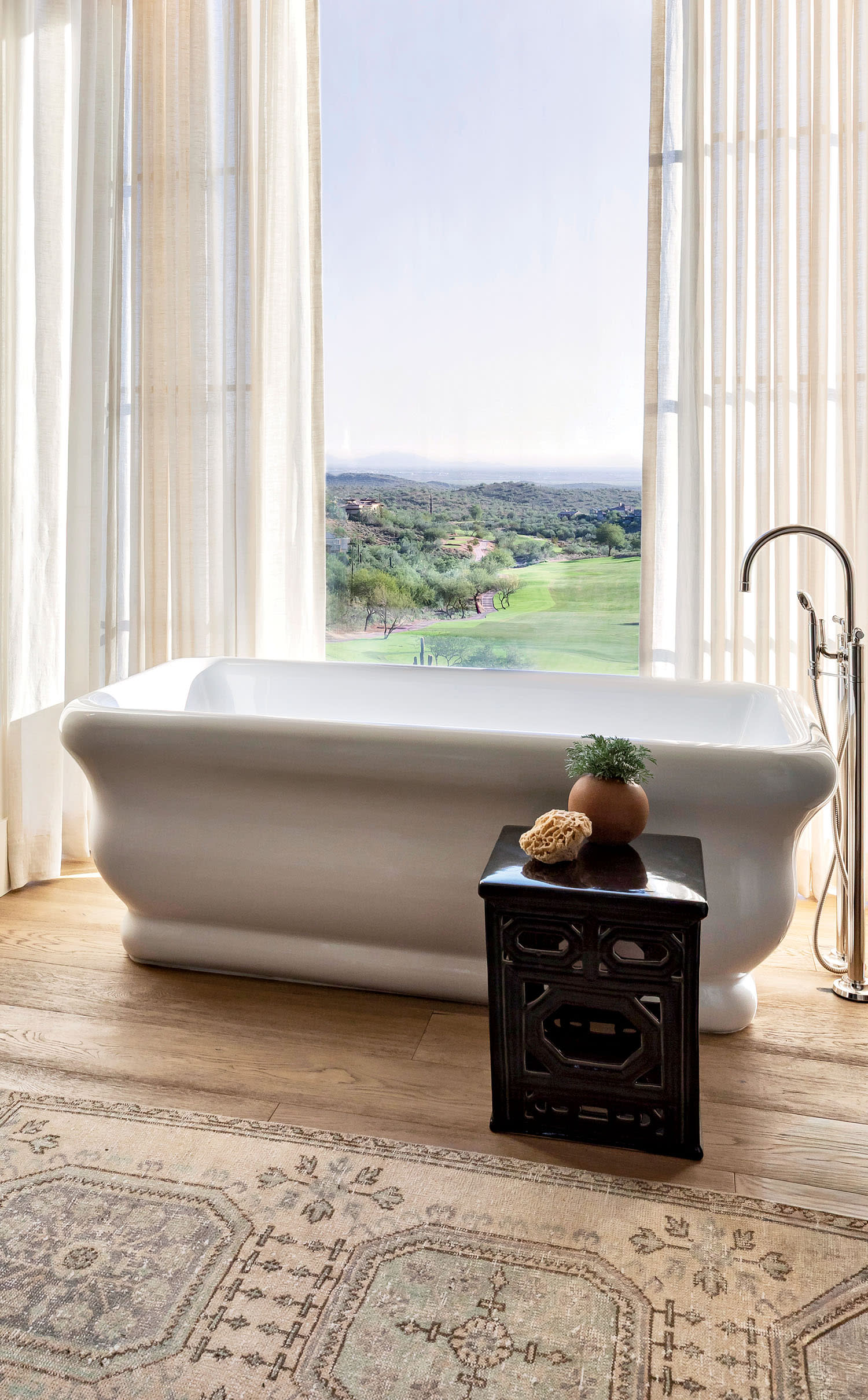 Luxurious bathroom with a freestanding white tub by a large window with sheer curtains, offering a scenic view. Wooden cabinet and plush rug add warmth.