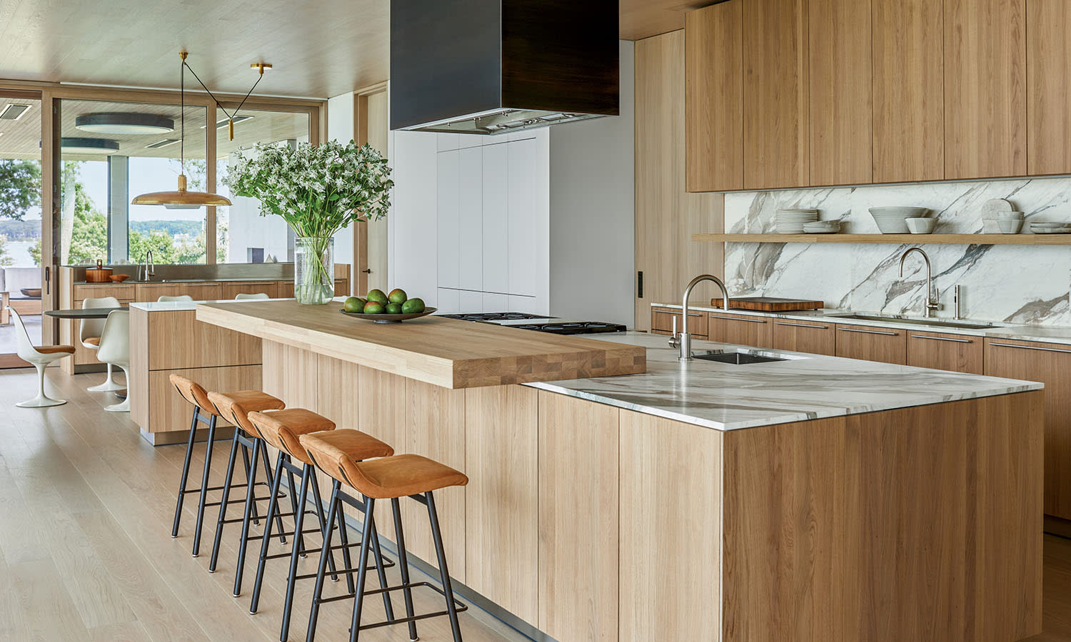 Modern kitchen with light wood cabinets and marble countertops. Features a large island with bar stools, a vase of white flowers, and a bowl of limes. 