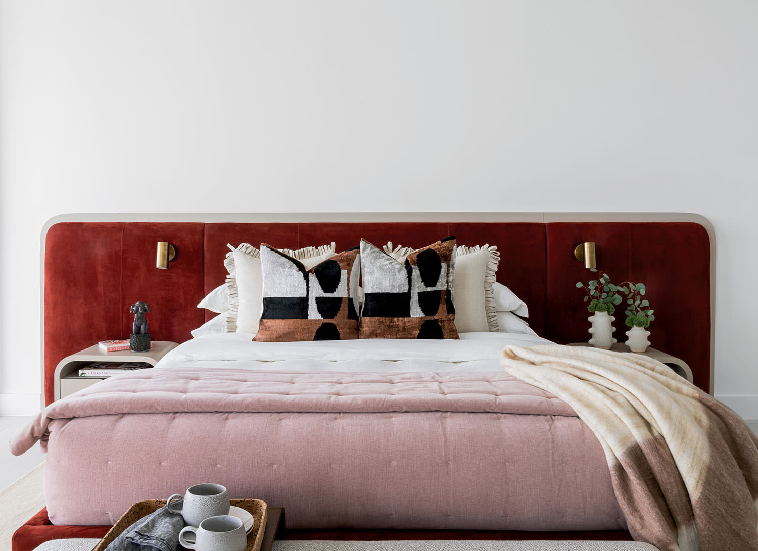 Luxurious bedroom with a large bed featuring a red headboard and patterned pillows. A pink bench at the foot holds a tray with cups. Modern, cozy vibe.