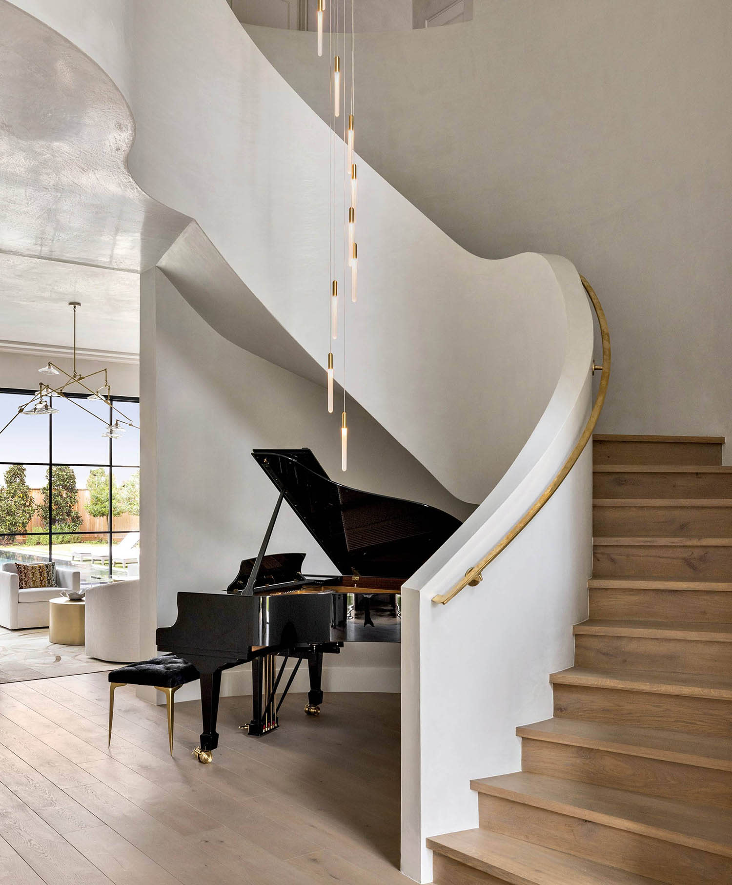 A paino sits beneath a curved staircase with white walls and a pendant light hanging from above. 