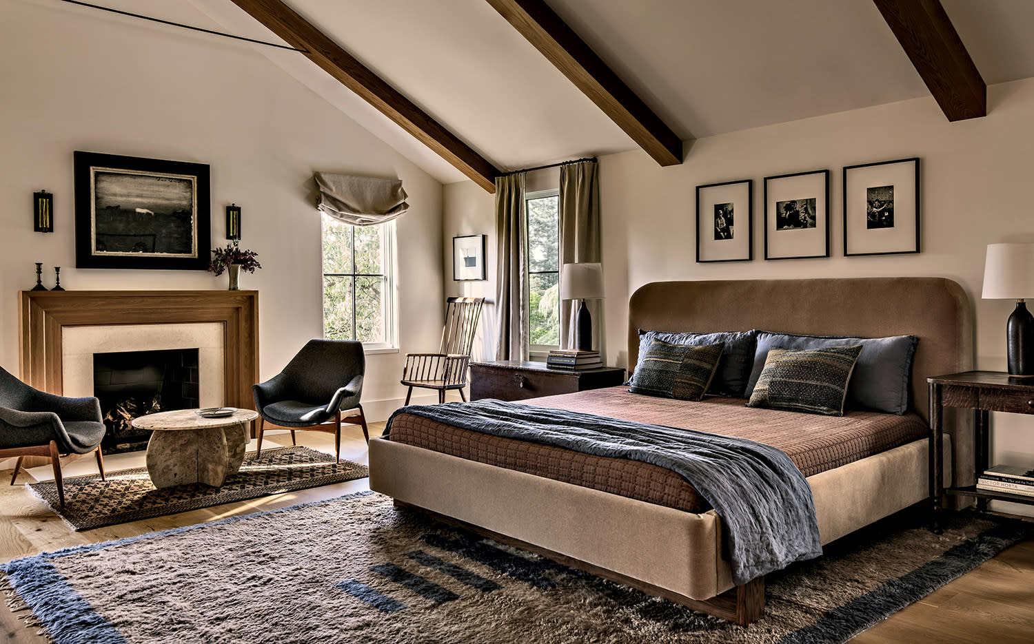 A cozy bedroom with exposed beams features a large bed, soft blankets, and pillows. A fireplace, chairs, and framed photos add warmth and style.